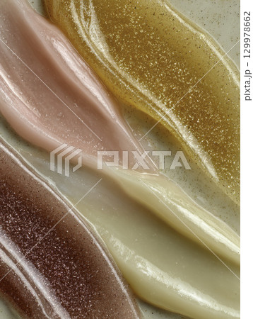 Macro closeup of various shimmering lip gloss swatches in pink, gold, red, and nude tones, showing glossy and glittery makeup textures 129978662
