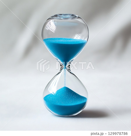 Hourglass with blue sand flowing down in front of white backdrop. 129978786