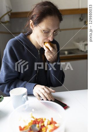 Woman eating fruit and using smartphone in kitchen Woman eating fruit and using smartphone in kitchen 129980033