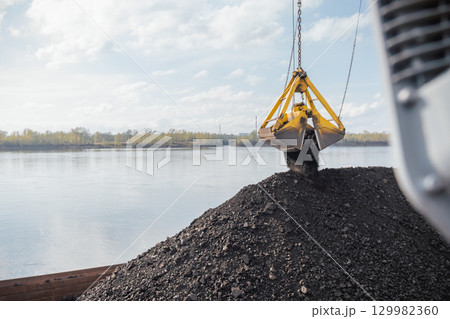 Coal is being loaded at the port using a crane over the river during a sunny day, Coal is poured onto a barge against the background of a river from a bucket Coal is being loaded at the port using a crane over the river during a sunny day, Coal is poured onto a barge against the background of a river from a bucket 129982360