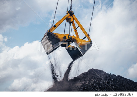 Heavy machinery is actively involved in unloading coal from a large bucket at an industrial location. The excavator crane, equipped with a dual clamshell bucket, hovers above a substantial pile of 129982367