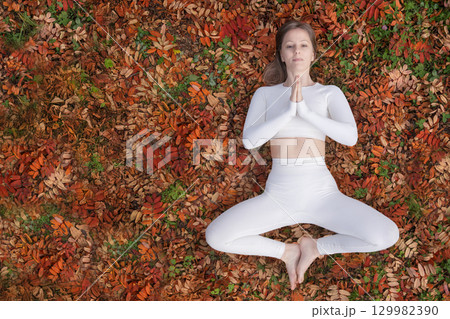 In peaceful outdoor setting during autumn, a woman performs a yoga pose on a vibrant carpet of colorful leaves. Dressed in white workout clothing, maintains meditative stance hands in prayer position In peaceful outdoor setting during autumn, a woman performs a yoga pose on a vibrant carpet of colorful leaves. Dressed in white workout clothing, maintains meditative stance hands in prayer position 129982390