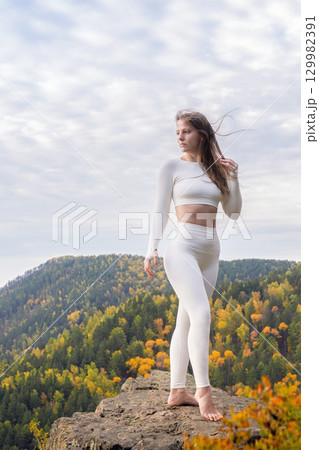 Athletic fitness woman poses in the distance on a stone cliff surrounded by mountains, autumn leaves. Playing sports and traveling outdoors 129982391