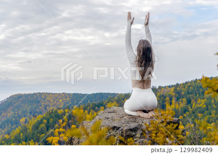 Woman sits on a rocky outcrop atop a mountain, engaged in yoga practice amidst vibrant autumn foliage. With her hands raised and back to the viewer, she embodies peace and focus in serene outdoors 129982409