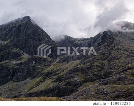 Carrauntoohil, the highest peak in Ireland, stands tall against a backdrop of moody clouds and steep, rocky slopes. The scene captures the wild beauty of Ireland's terrain. Carrauntoohil, the highest peak in Ireland, stands tall against a backdrop of moody clouds and steep, rocky slopes. The scene captures the wild beauty of Ireland's terrain. 129983289