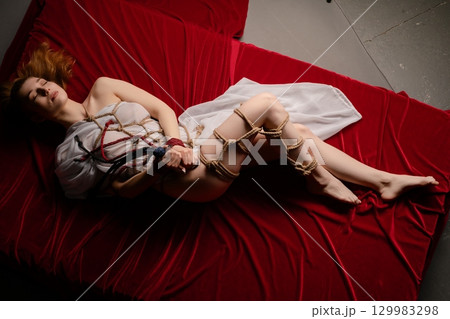 Exploring artistic shibari techniques with a bound woman on a red platform Exploring artistic shibari techniques with a bound woman on a red platform 129983298