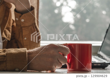 person holding red mug while working on laptop near window cozy soft natural light. closeup. person holding red mug while working on laptop near window cozy soft natural light. closeup. 129984629