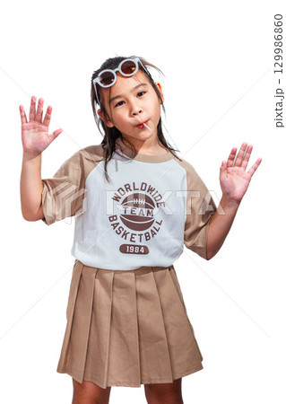 An Asian girl with sunglasses on her head eating a lollipop and raising both hands, posing playfully with a fun expression against a white background. 129986860