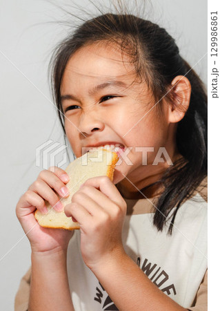 An Asian girl smiling while biting into a slice of bread, showing a playful and happy expression during mealtime. 129986861
