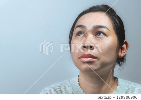 An Asian woman wearing a light sweater looking upward with a thoughtful and slightly worried expression against a grey background. An Asian woman wearing a light sweater looking upward with a thoughtful and slightly worried expression against a grey background. 129986906