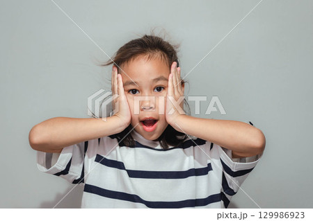 A surprised Asian girl with her hands on her cheeks and mouth open, wearing a striped shirt and showing an amazed expression. A surprised Asian girl with her hands on her cheeks and mouth open, wearing a striped shirt and showing an amazed expression. 129986923