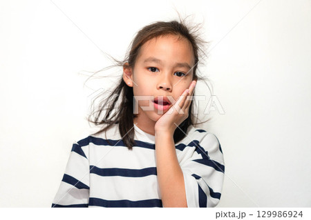 A surprised Asian girl touching her cheek with one hand, wearing a striped shirt and showing an amazed expression against a white background. 129986924