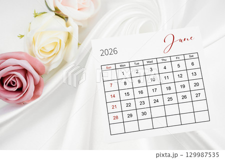 June 2026 calendar page and rose flower on white satin textile background. 129987535