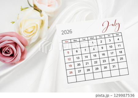 July 2026 calendar page and rose flower on white satin textile background. July 2026 calendar page and rose flower on white satin textile background. 129987536
