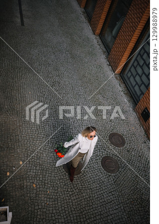 Woman walking on cobblestone street holding flowers in urban setting during day 129988979