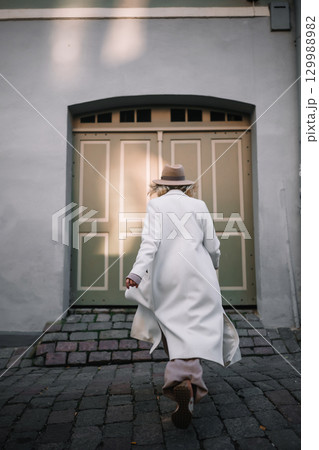 Woman in white coat with hat walking toward vintage door on cobblestone street 129988982