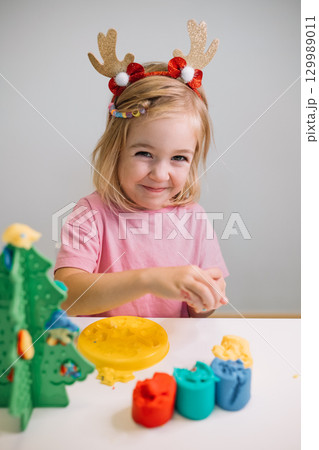 A young girl wearing festive antlers enjoys crafting holiday decorations at a table 129989011