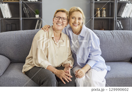 Cheerful adult woman and her elderly mother posing together on sofa, embracing warmly 129990602