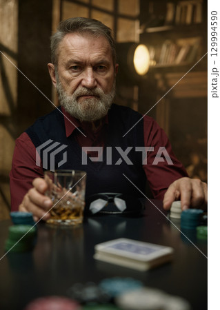 An experienced mature man sits at the poker table enjoying his whiskey and playing with chips An experienced mature man sits at the poker table enjoying his whiskey and playing with chips 129994590