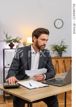 Middle-aged businessman accountant enters numbers on laptop pc checks receipts with calculator signs 129996736
