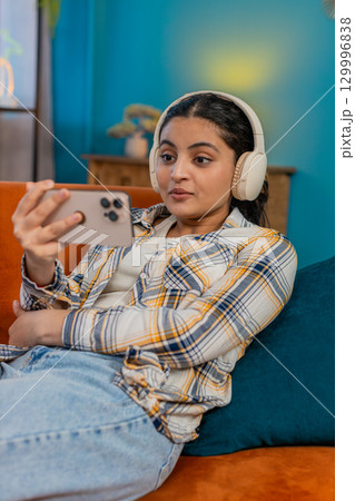 Indian woman relaxing on sofa wearing headphones happily listening podcast audiobook from smartphone Indian woman relaxing on sofa wearing headphones happily listening podcast audiobook from smartphone 129996838