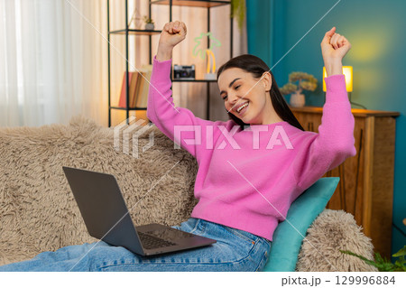Young woman on sofa raising hands up joyfully celebrating successful finished project on laptop 129996884