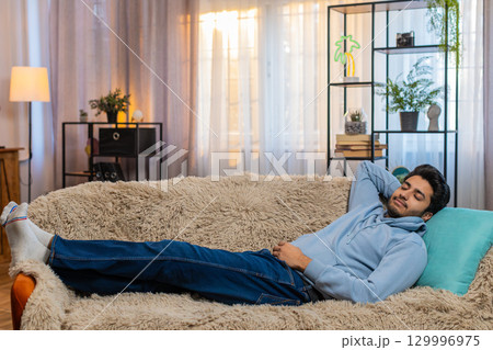 Tired Indian man lying on comfortable couch taking rest and daytime nap in living room at home Tired Indian man lying on comfortable couch taking rest and daytime nap in living room at home 129996975