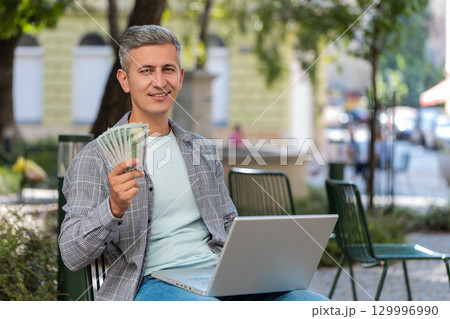 Happy rich man using laptop winner victory holding cash money in dollar banknotes sitting on chair 129996990