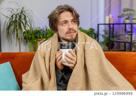 Sick ill Caucasian man wrapped in plaid sit shivering from cold drinking hot coffee or tea at home 129996999