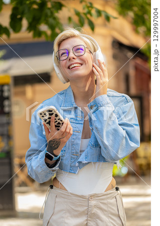 Overjoyed blonde woman in wireless headphones choosing, listening music holding smartphone on street 129997004