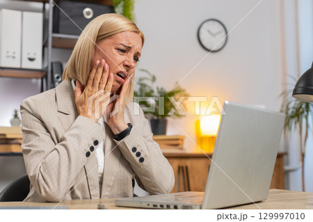 Annoyed mature businesswoman looking at laptop screen irritated working remotely at office home 129997010