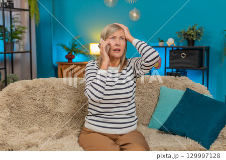 Mature woman looking upset anxious while listening to bad news on mobile phone sitting on home sofa 129997128