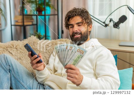 Successful rich young Indian man holding dollar bill and smartphone sitting on sofa couch at home 129997132