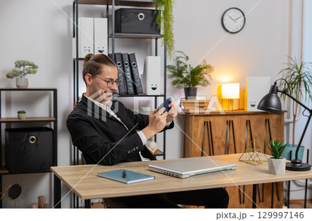 Excited young businessman playing shooter or racing video games on smartphone sitting at desk Excited young businessman playing shooter or racing video games on smartphone sitting at desk 129997146