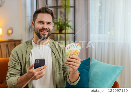 Successful rich young Caucasian man holding euro bill money fan sitting on sofa couch at home 129997150