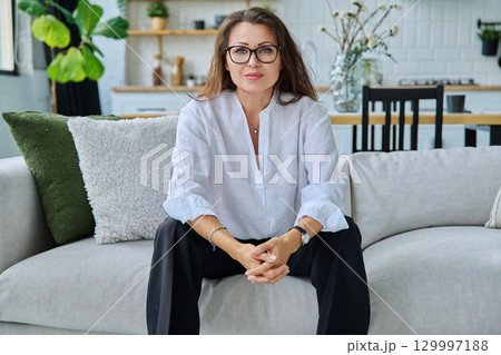 Successful beautiful mature woman looking at camera sitting at home on sofa 129997188