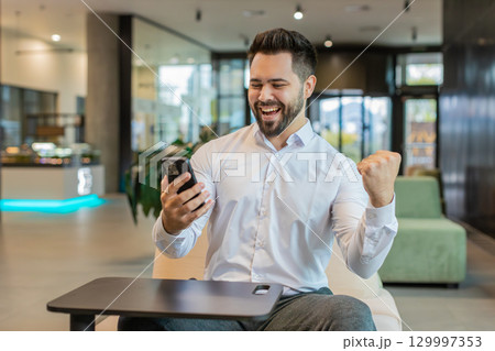 Happy young businessman using smartphone shocked by sudden victory lottery win in office lobby 129997353