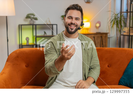 Confident young Caucasian man guy sitting on sofa at home wearing headset talking with client Confident young Caucasian man guy sitting on sofa at home wearing headset talking with client 129997355