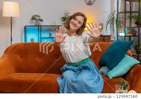Happy young woman vlogger smiling friendly at camera and waving hands while sitting on sofa at home Happy young woman vlogger smiling friendly at camera and waving hands while sitting on sofa at home 129997360