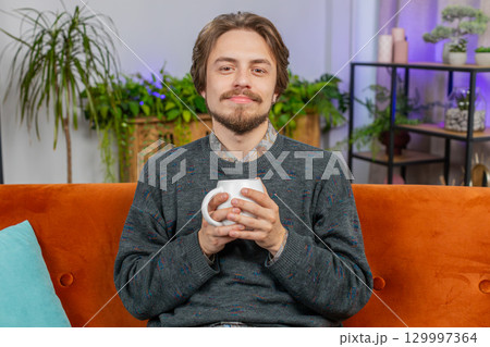 Handsome smiling Caucasian young man drinking a cup of coffee or herbal tea sitting at home couch 129997364