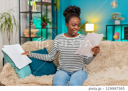 African American woman upset stressed with financial problems sitting at home, looking at documents 129997365