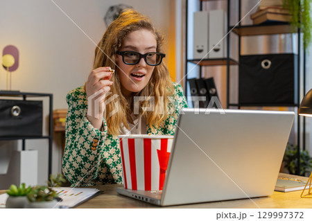 Businesswoman taking break from work wearing 3D glasses eating popcorn and watching movie at office Businesswoman taking break from work wearing 3D glasses eating popcorn and watching movie at office 129997372