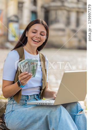 Happy rich woman using laptop winner victory holding cash money in dollar banknotes on city street Happy rich woman using laptop winner victory holding cash money in dollar banknotes on city street 129997379