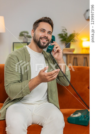 Smiling young man making wired telephone call conversation with friends sitting on sofa at home Smiling young man making wired telephone call conversation with friends sitting on sofa at home 129997396