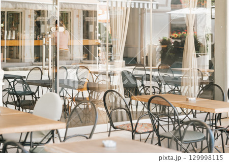 Cozy outdoor cafe with empty tables and chairs seen through reflective glass window. Concept of summer leisure, end of season, peaceful urban relaxation Cozy outdoor cafe with empty tables and chairs seen through reflective glass window. Concept of summer leisure, end of season, peaceful urban relaxation 129998115