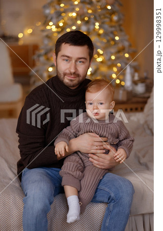 Father holding baby son in cozy room with Christmas tree, wearing warm knitted sweaters 129998351