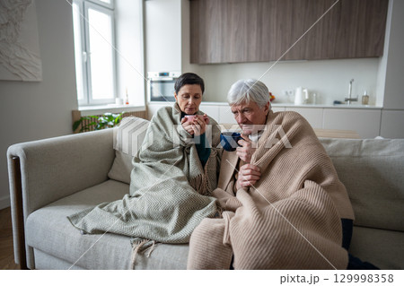 Upset struggling senior couple under blanket sipping hot tea remedy, suffer from illness at home 129998358