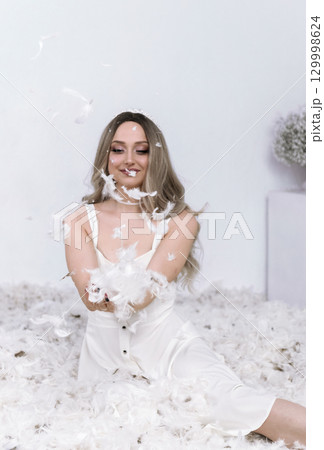 a very beautiful blonde sits in a white room in which feathers are flying 129998624