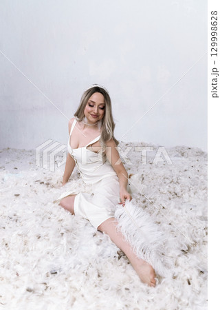 a very beautiful blonde sits in a white room in which feathers are flying 129998628