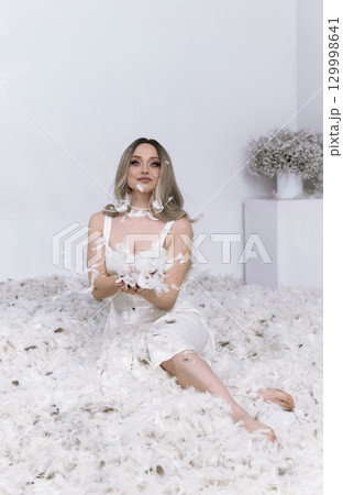 a very beautiful blonde sits in a white room in which feathers are flying 129998641
